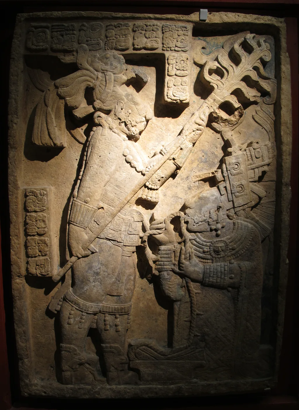 A Maya carved lintel shows the kneeling Lady K'ab'al Xook drawing a rope through her tongue while a standing ruler holds a torch above her.